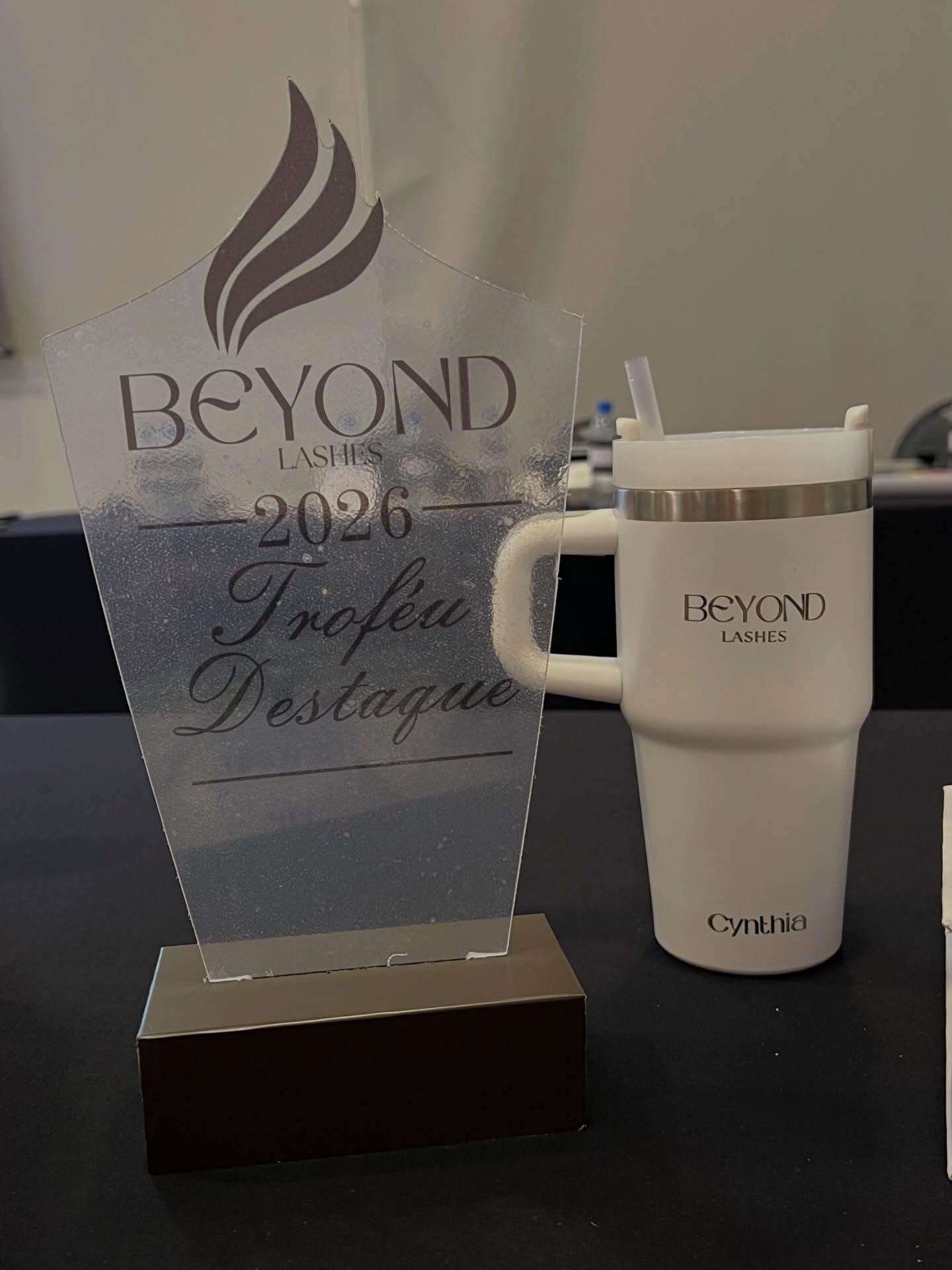 Troféu de acrílico gravado BEYOND LASHES 2026, Troféu Destaque, ao lado do copo térmico branco com o nome Cynthia.